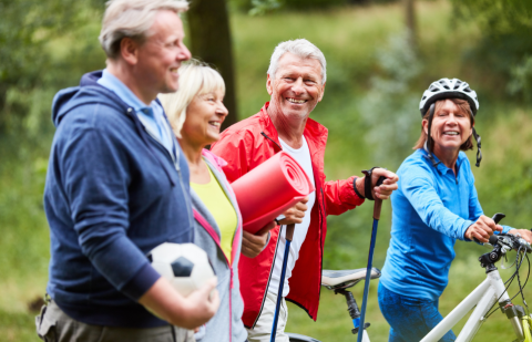 Grupa seniorów na spacerze trzymająca sprzęt sportowy: piłkę, matę, kijki, rower.